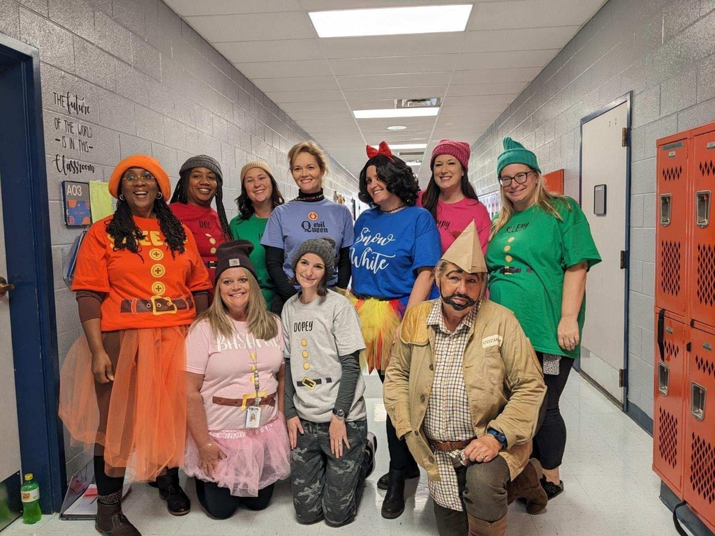 Nine people in costumes pose for a photo in a school hallway. They stand in two rows, wearing colorful costumes.
