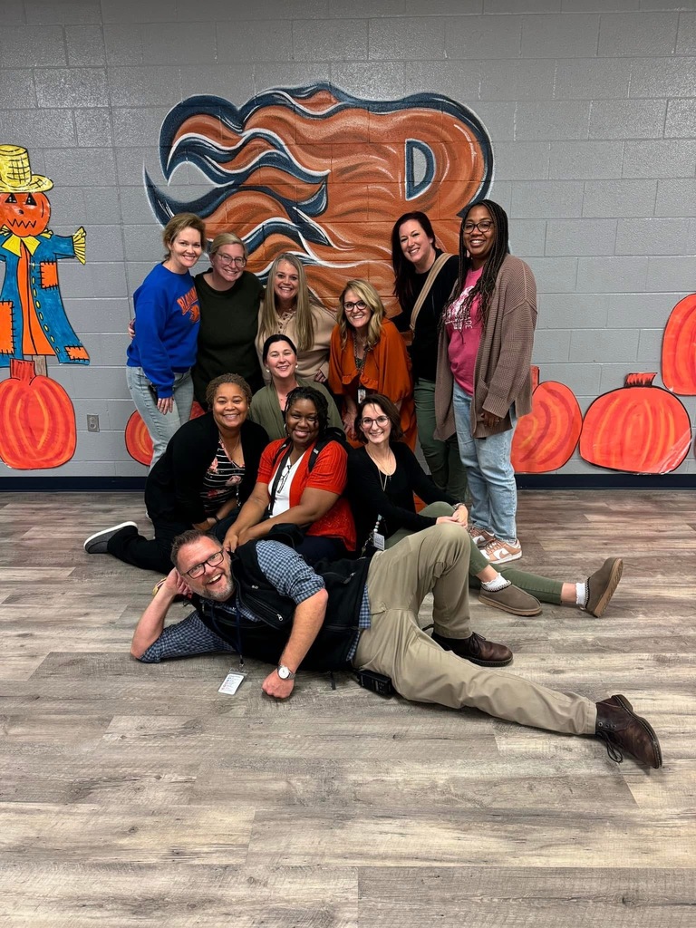 Alt Text:  Group photo of eleven Blackman Elementary staff members posed in front of a large orange-and-blue “B” mural decorated with flame accents. The group is arranged in several rows, with some seated or kneeling on the floor and one staff member playfully reclining in the front. Fall-themed decorations, including painted pumpkins and a scarecrow, line the wall around the mural. Staff members wear a mix of school spirit wear and casual professional attire in fall colors. The photo was taken indoors on a wood-look floor with a gray cinderblock wall backdrop.