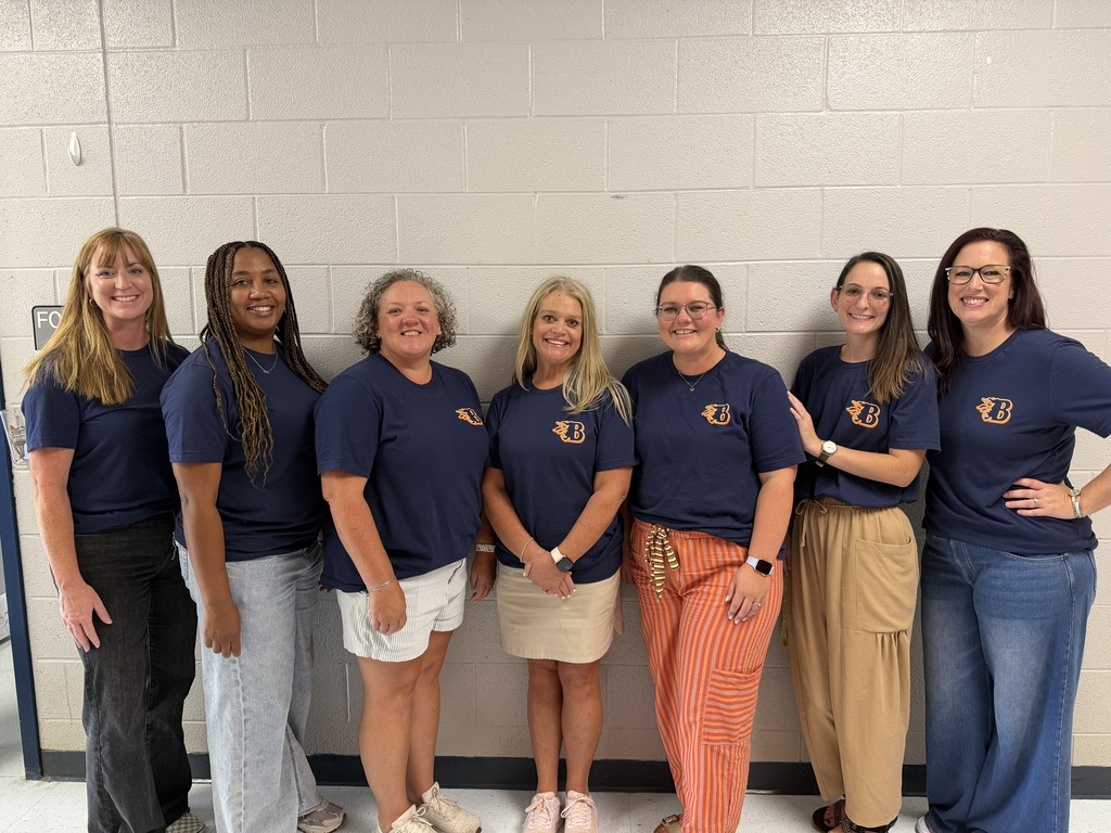 Alt Text: Seven Blackman Elementary staff members stand side by side in a school hallway against a light-colored cinderblock wall. Each person is wearing a navy blue Blackman Elementary T-shirt with an orange and blue “B” logo on the front. The group is dressed in a variety of casual and professional attire, including jeans, skirts, shorts, and patterned pants. The photo was taken indoors with bright lighting, and the staff members are posed together for a group picture.