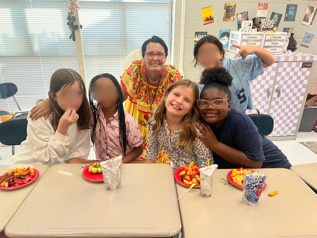 A group of students and a teacher seated at a table with plates of food. The classroom has chairs, posters, and a refrigerator.