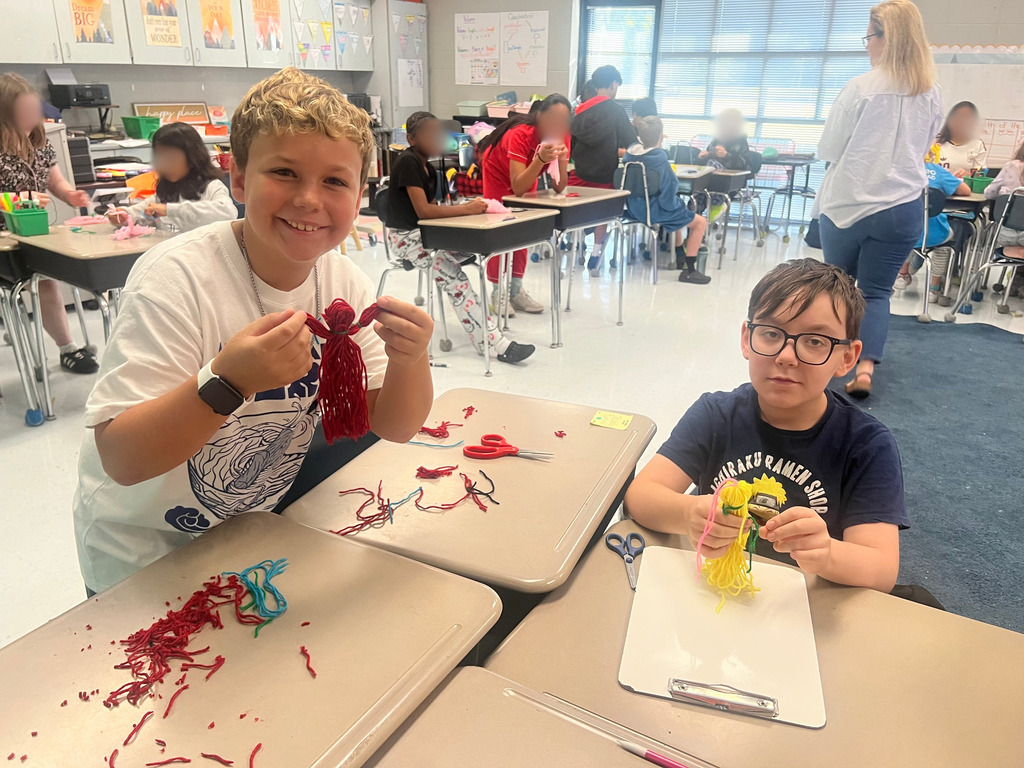 Two children at desks in a classroom, one holding a red craft material, the other using scissors on a project.