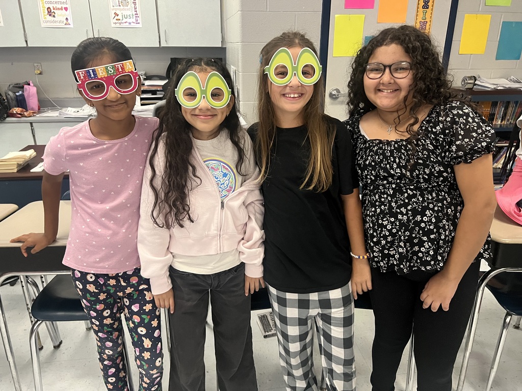 Four girls in a classroom wearing identical masks, standing together. A woman in the background wears glasses and a black floral shirt.