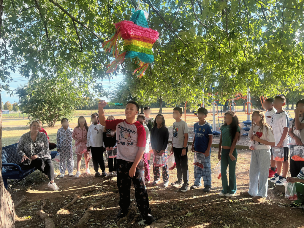 A group of children and adults gather under a tree with a hanging pinata, looking on.