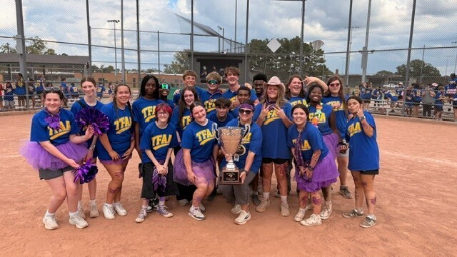 A sports team stands on a dirt field with a fence in the background, holding a trophy.