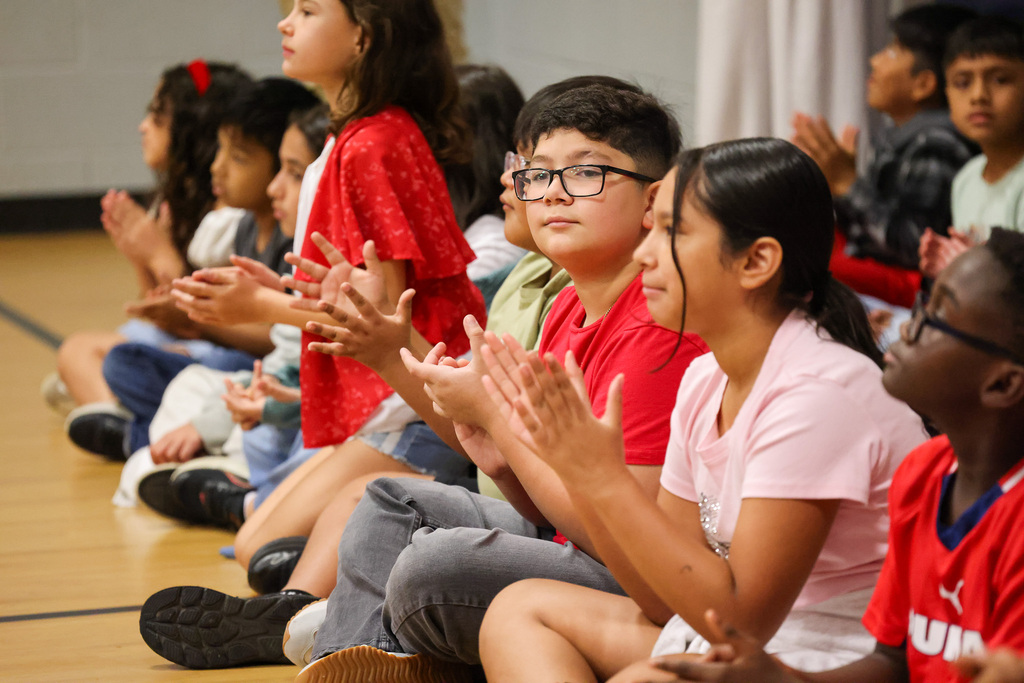 Group of children clapping hands seated on a wooden floor in a school gym.