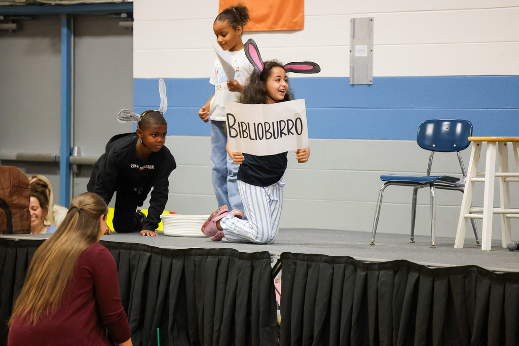 Children on stage, one holding a sign, another kneeling, one standing with bunny ears. Audience in background.