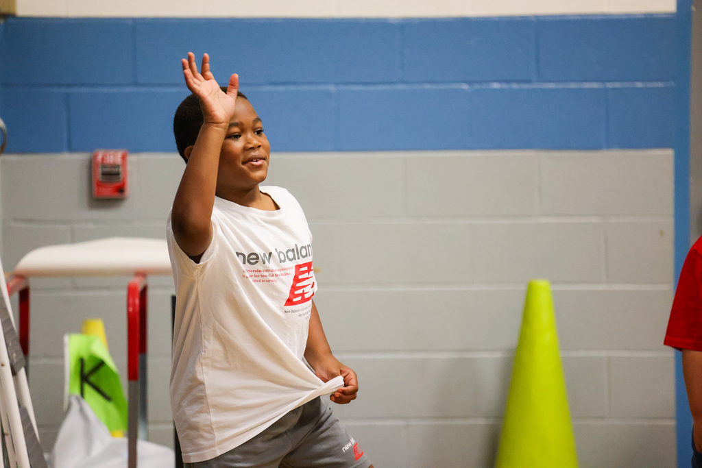A child in a white t-shirt waves enthusiastically in an indoor sports area with blue and white walls.