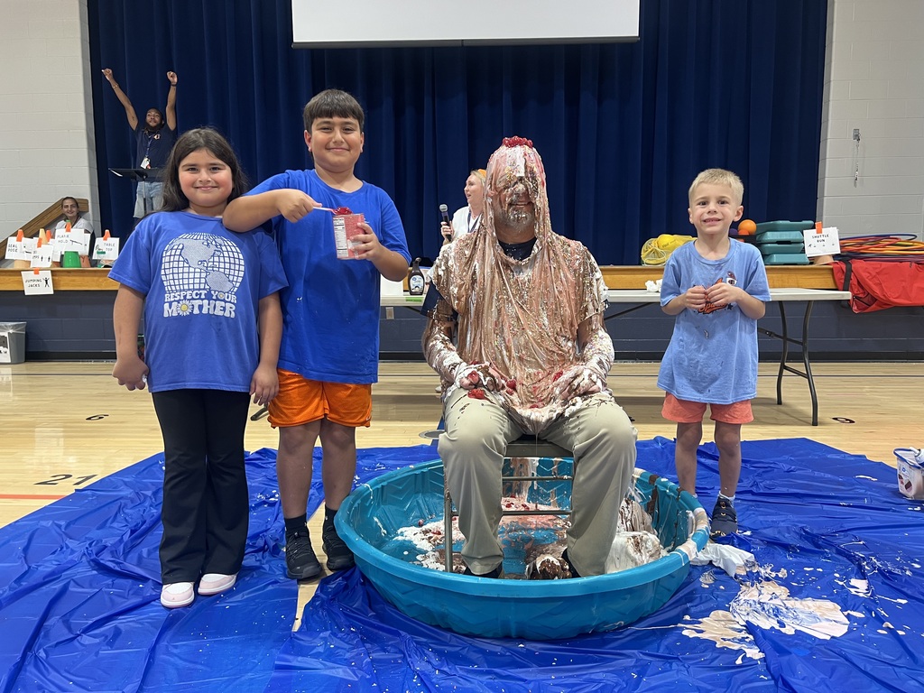Four children stand around a blue pool on a gym floor. A man sits in the pool with a splattered face.