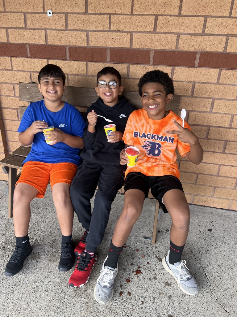 Three boys in shorts and sneakers sitting on a bench, holding cups of ice cream, with brick wall in background.