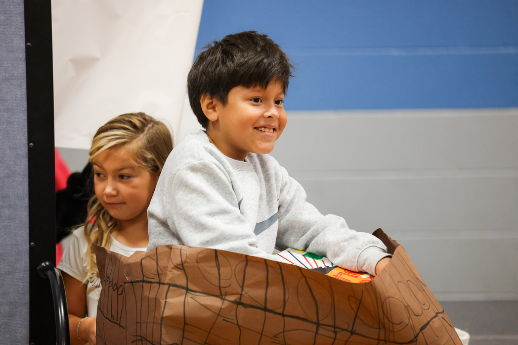 A boy and girl sit together, smiling, with the boy holding a bag. The bag is brown and patterned.