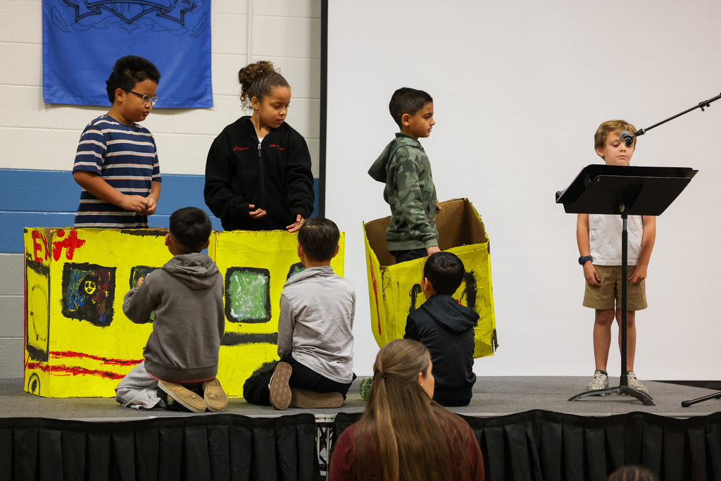 Children in costumes stand on stage, some sitting in cardboard boxes, in a classroom setting.