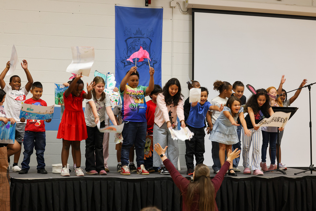 A group of children stand on stage, some raising their hands, holding papers, and performing. A banner is visible behind them.
