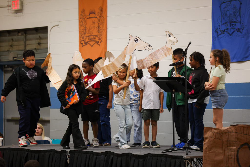 Children stand on stage with paper camels, some holding them, others not. A podium and microphone are present.