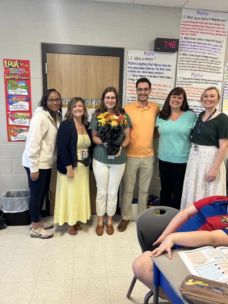 A group of people stand together in a classroom. Some hold flowers. A trash can is near the door.