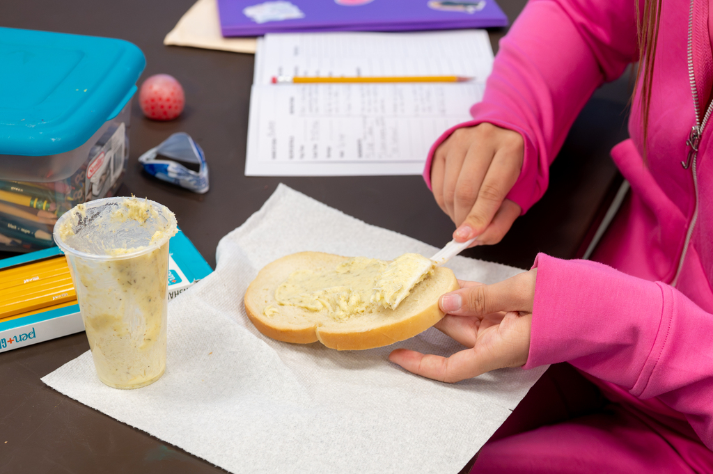 Person in pink top spreads white spread on sandwich on paper. A cup of yellow liquid is nearby.
