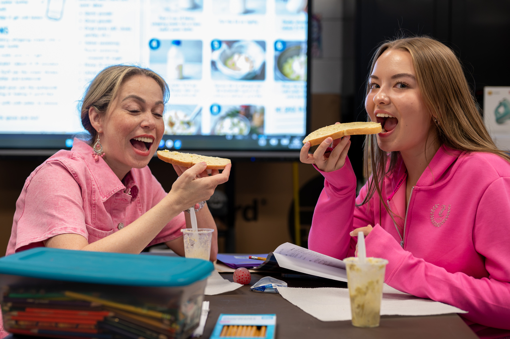 Two women in pink shirts seated, each holding a sandwich. A table has paper, a plastic container, and drinks. A screen in background displays images.