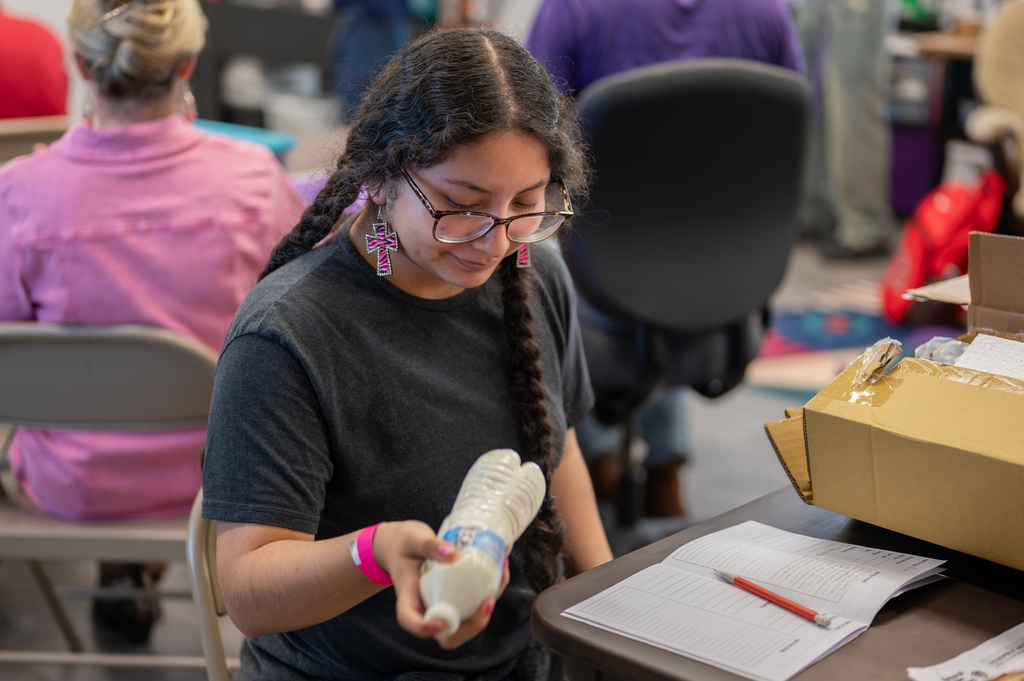 A young woman examines a prosthetic hand while sitting at a desk. Other people are seated nearby.