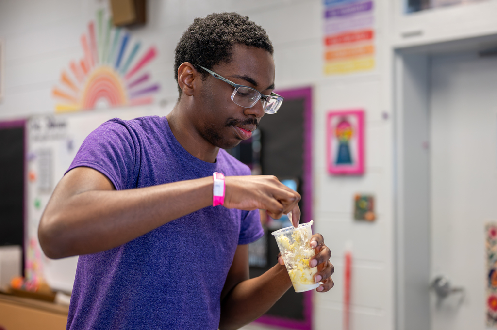 A person in a purple shirt and glasses stirs a drink in a plastic cup. Bright colors decorate the classroom walls.