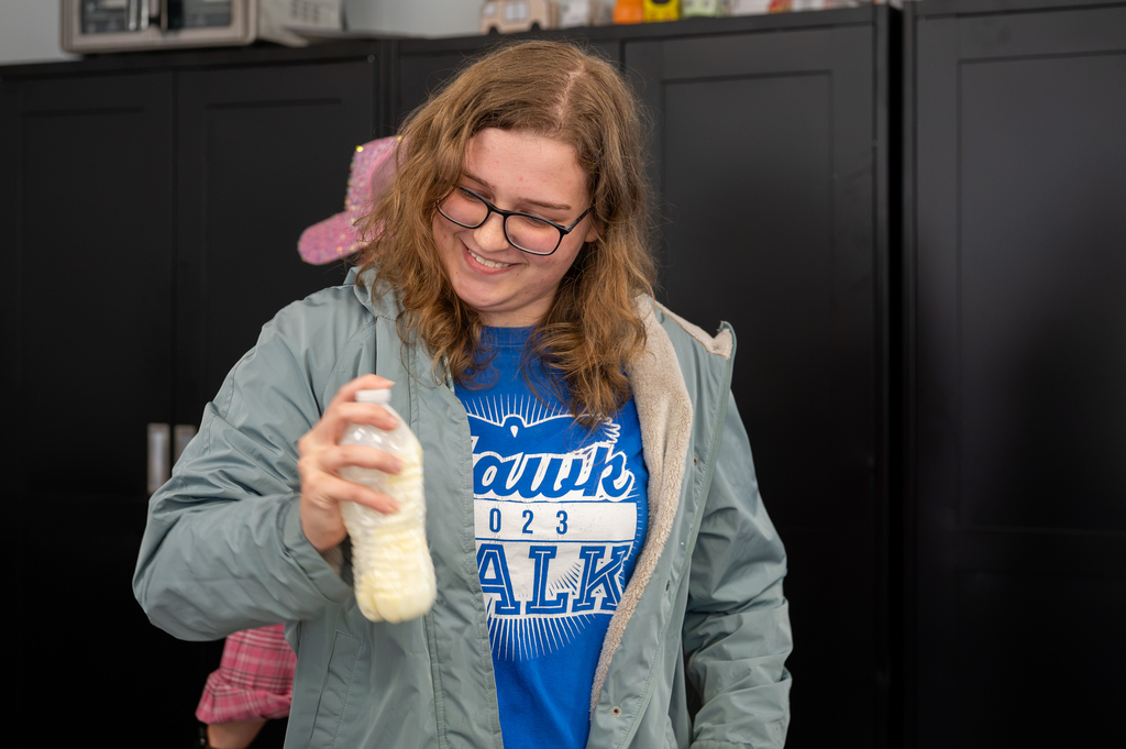 Woman holding a bottle of milk, wearing glasses, a blue shirt, and a light-blue jacket.