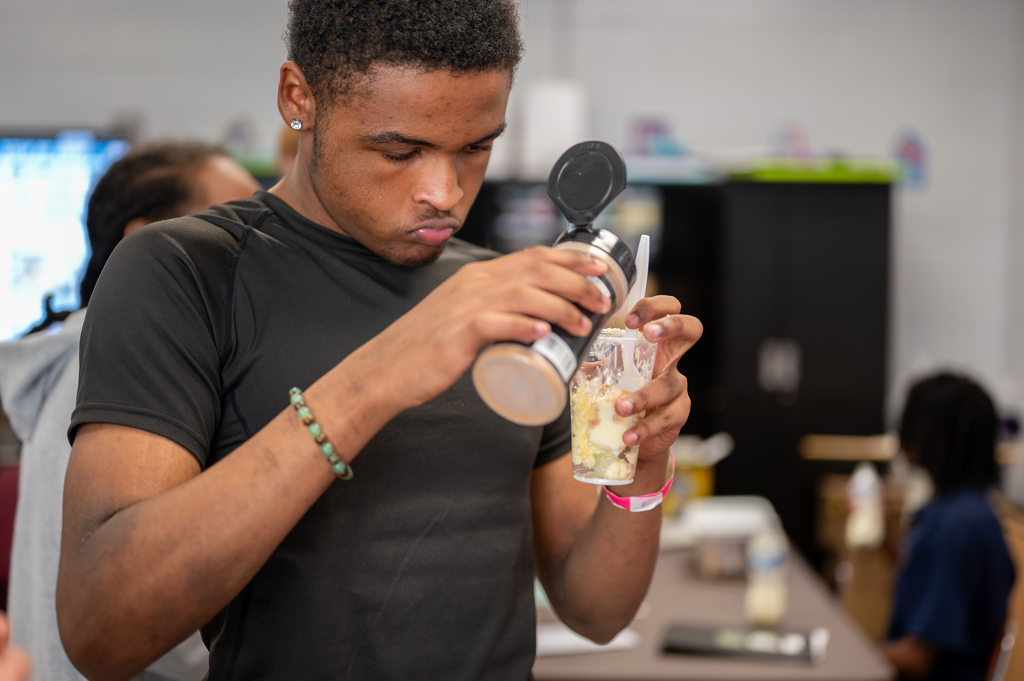 A man pours a smoothie into a cup in a kitchen. Behind him, others are seated at a table.