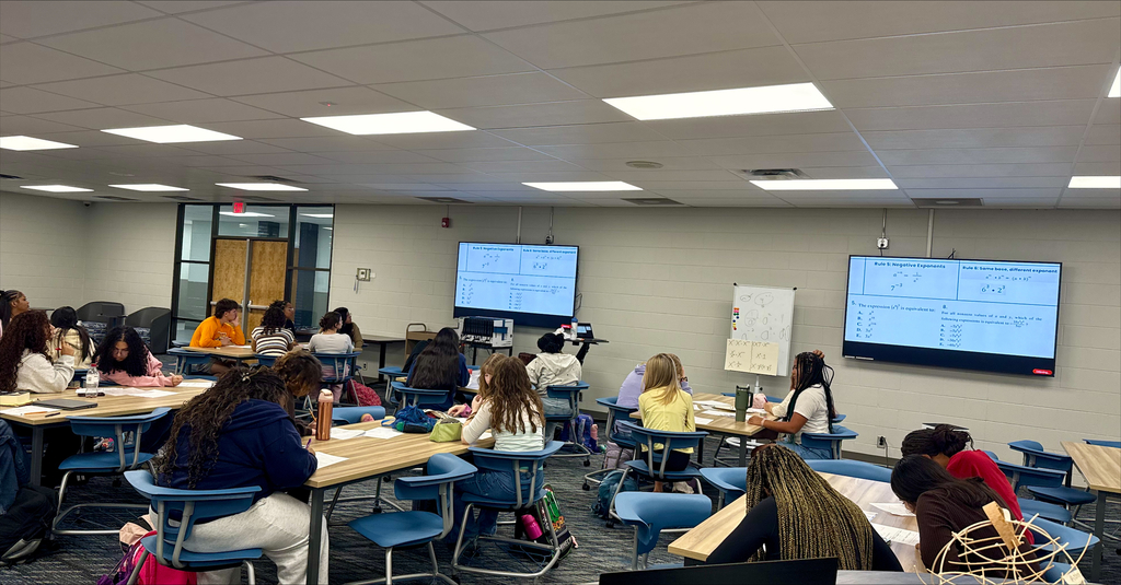 A classroom with several tables and chairs. Students sit at tables and work on papers. Monitors on the wall display information.