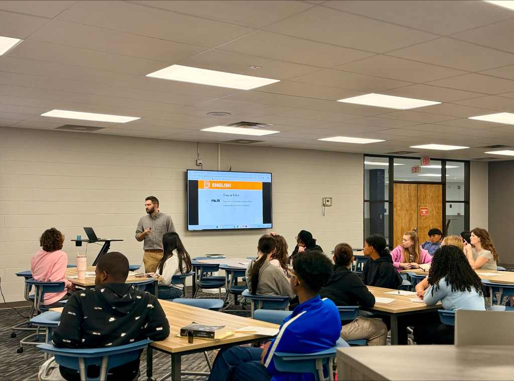 A classroom with students seated at tables, a man stands at a podium in front of a projector screen.