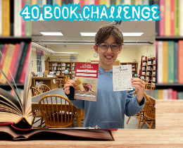 A boy wearing glasses holds a book and a paper inside a library. The background shows shelves of books and a table.