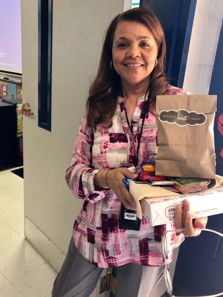 Woman holding a box and bag, smiling, standing in a hallway. She wears a pink and gray checkered shirt.