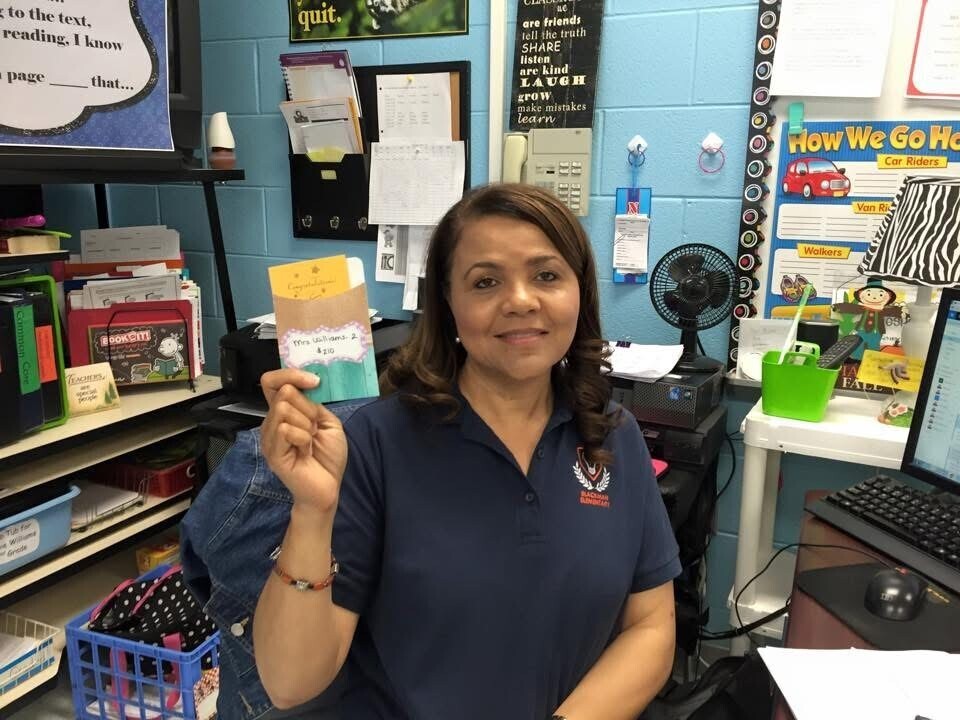 Woman wearing a navy blue shirt holding a book up to the camera, smiling. Behind her is a classroom with shelves of books, a computer, and a fan.