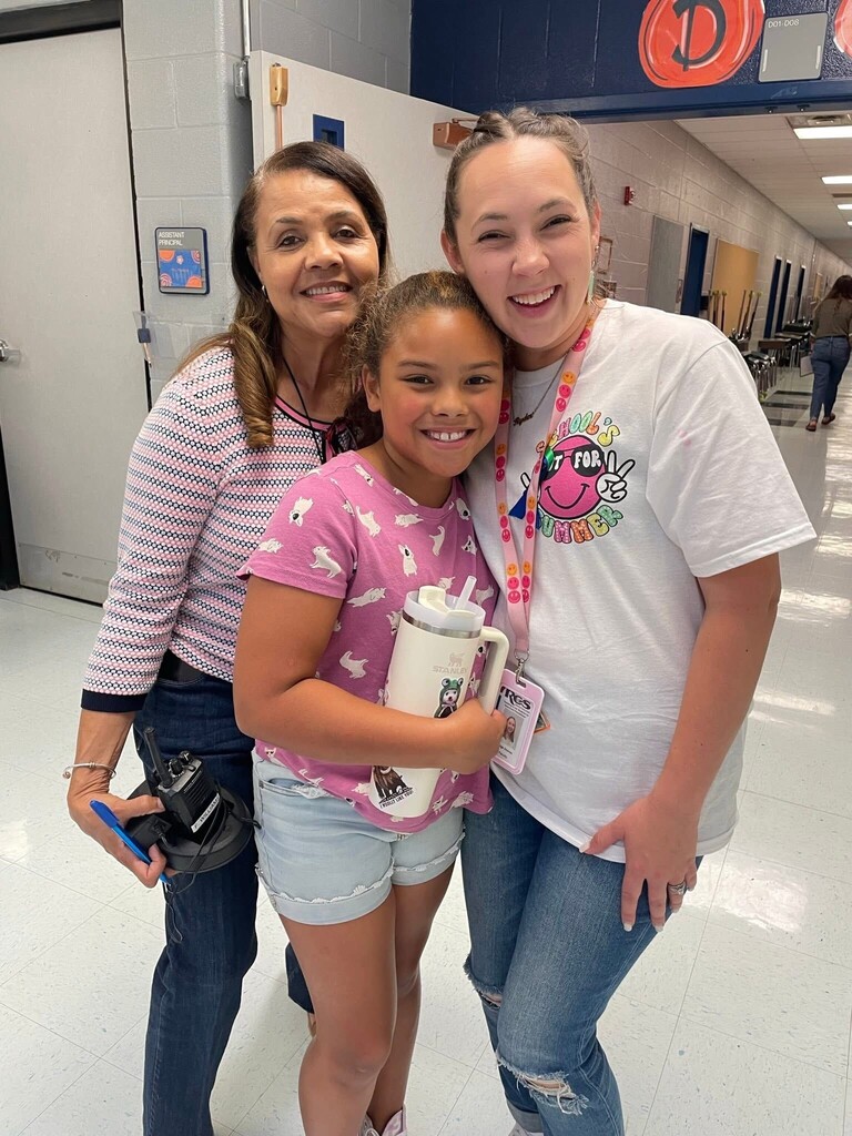 Three people stand in a hallway, smiling and posing for a photo. The left person holds a camera. Middle holds a cup. Right wears a white shirt and necklace.