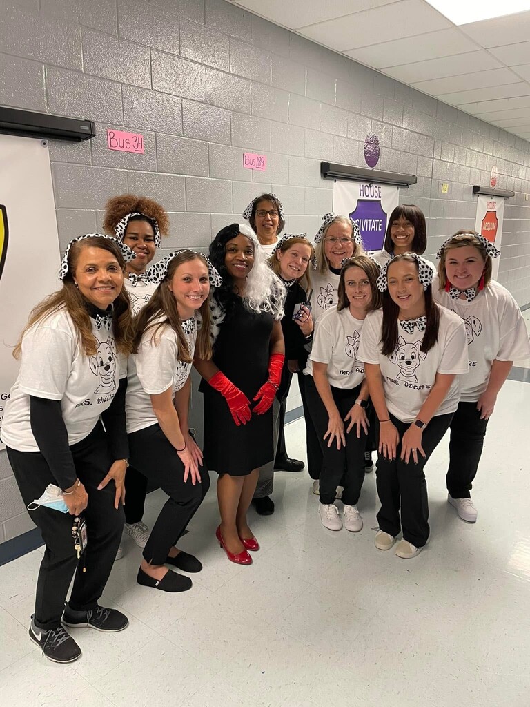 A group of women in matching white shirts and black pants, some wearing red gloves, stand in a hallway.