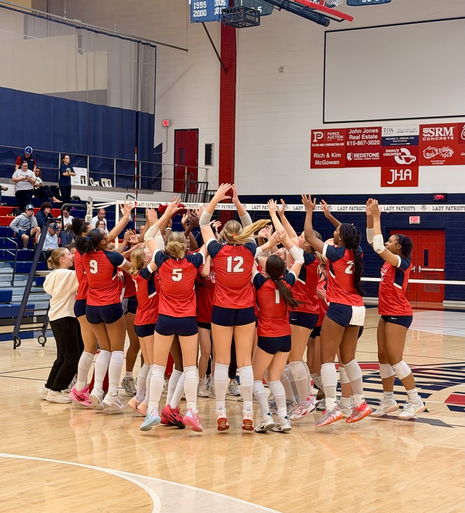 Volleyball players in red uniforms with numbers gather in a gym, forming a circle with hands raised.