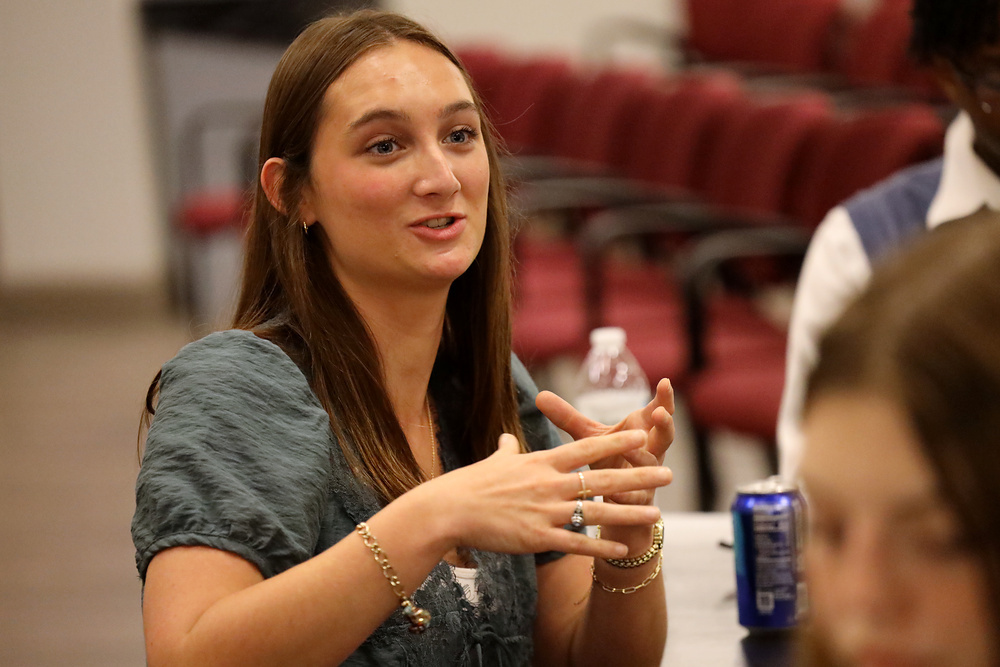 A woman wearing a sleeveless shirt speaks while gesturing with her hands in a room with red chairs.