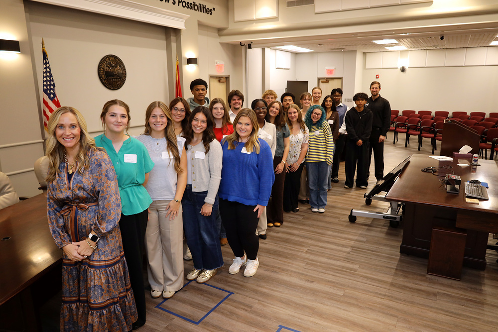 A group of adults and students standing in a room with a flag, wooden floors, and a table.