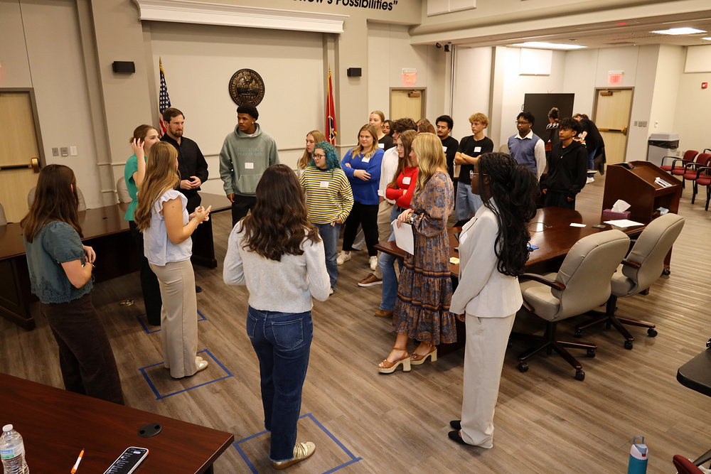 A group of students in a meeting room, standing in circles. Some people are talking.
