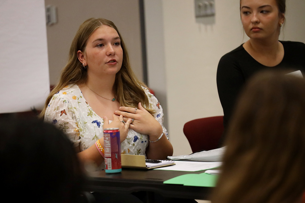 Three students in a classroom, one speaking with hands clasped, another listening with a cup nearby.