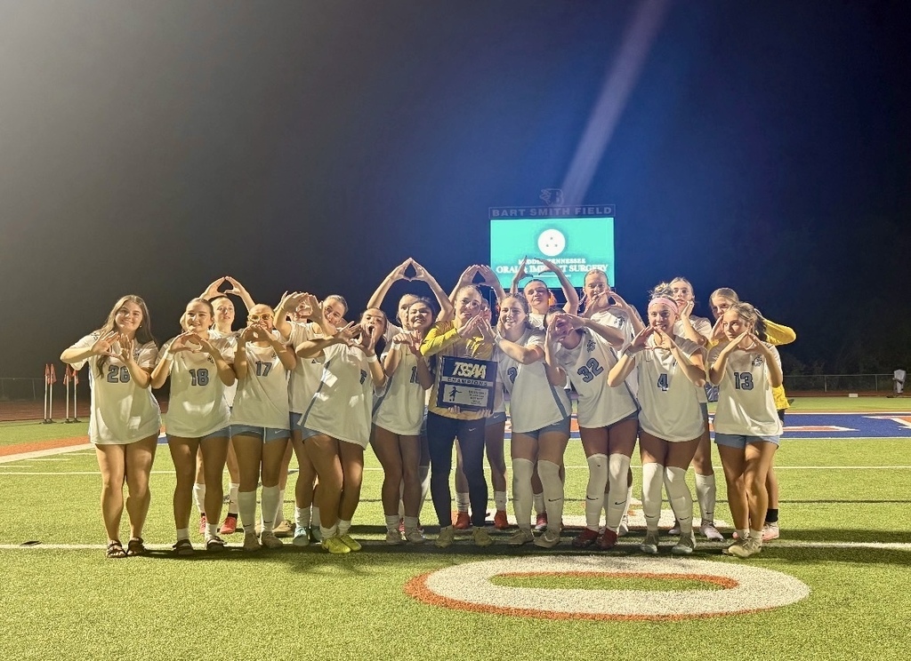 A sports team in white uniforms poses with a trophy on a field with a scoreboard.