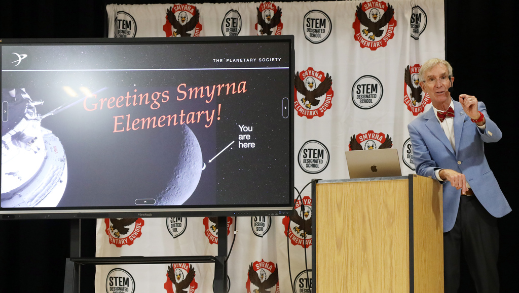 Person at podium speaking with a large screen showing a message and a planet behind.