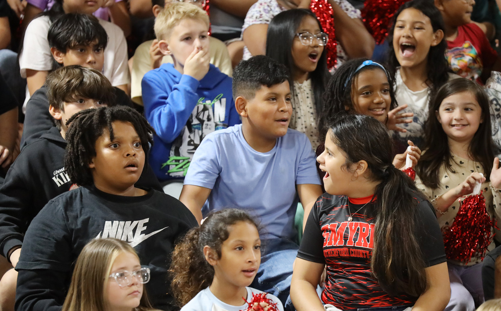 Diverse group of students sitting in bleachers, some wearing uniforms, others in casual attire, engaging in conversation.