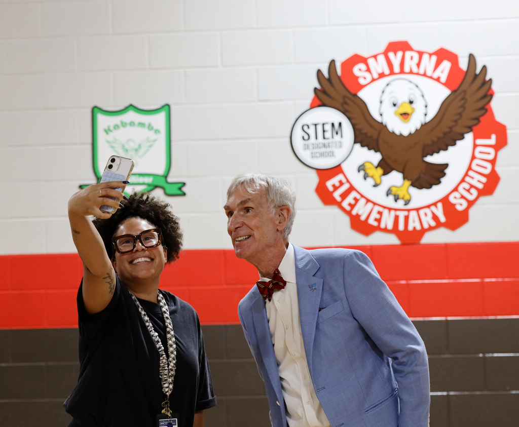 Two people, a woman taking a photo and a man in a bow tie, stand together against a wall with two logos.