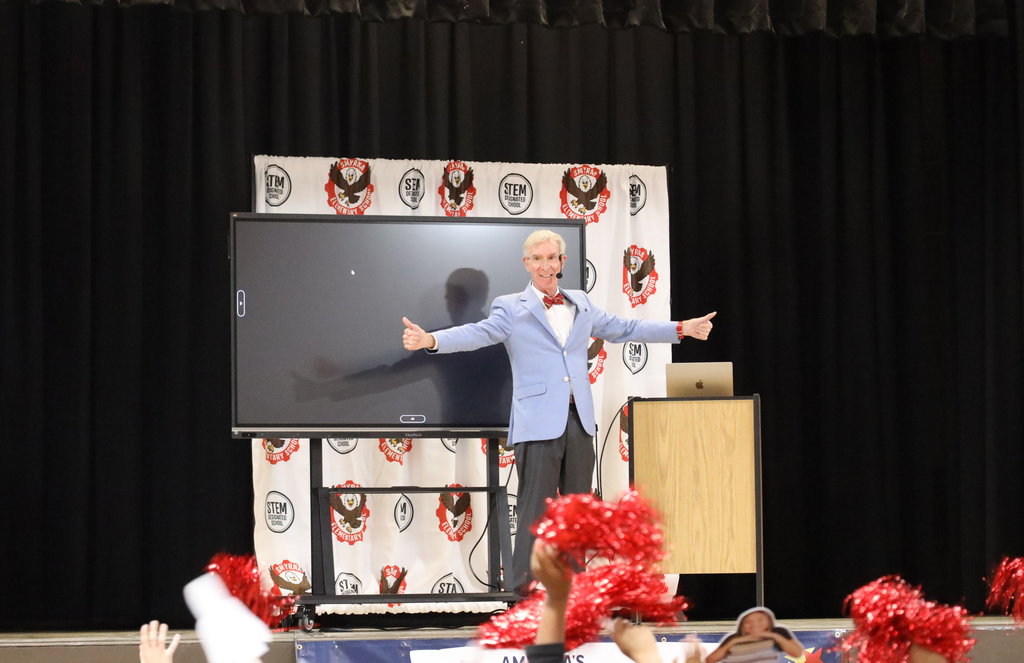 Man on stage with arms raised in front of large screen. Some students are raising their hands.