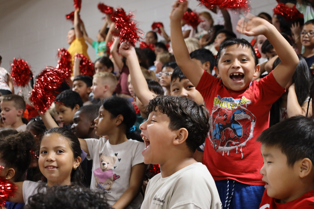 Many children are cheering and smiling with red pom poms and T-shirts. They are in a gymnasium.