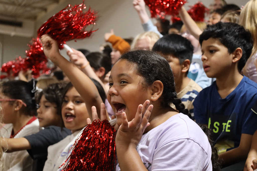 Students at a school event, holding red pom-poms, cheering with hands in the air.