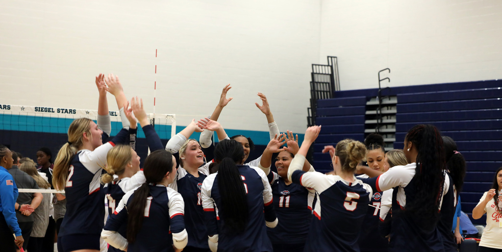 A group of young women, wearing matching blue and white uniforms, celebrate in a gymnasium.