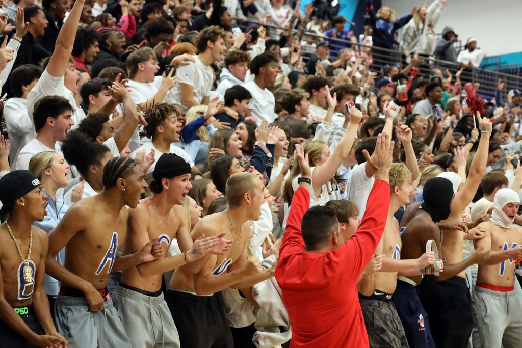 A crowd of students wearing sports jerseys cheer and raise hands in a gymnasium.