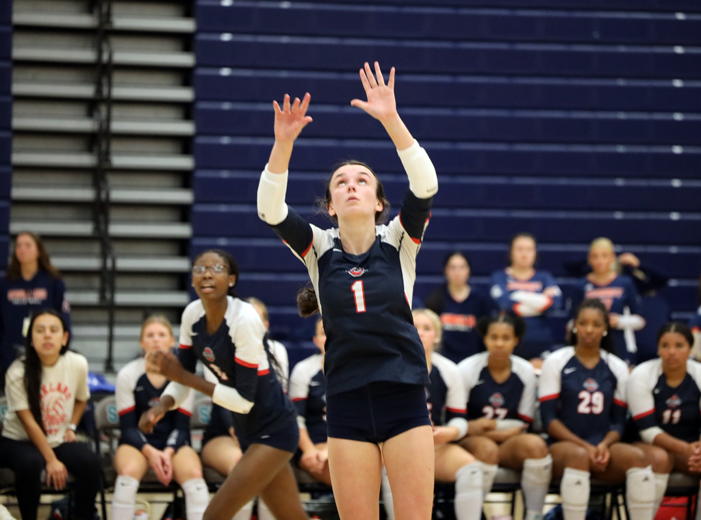 A girl in a white and blue uniform prepares to serve a volleyball. Other players sit behind her.