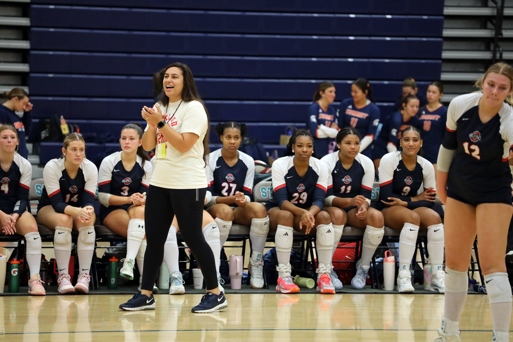 A woman speaks with hands raised in front of a seated group of volleyball players.