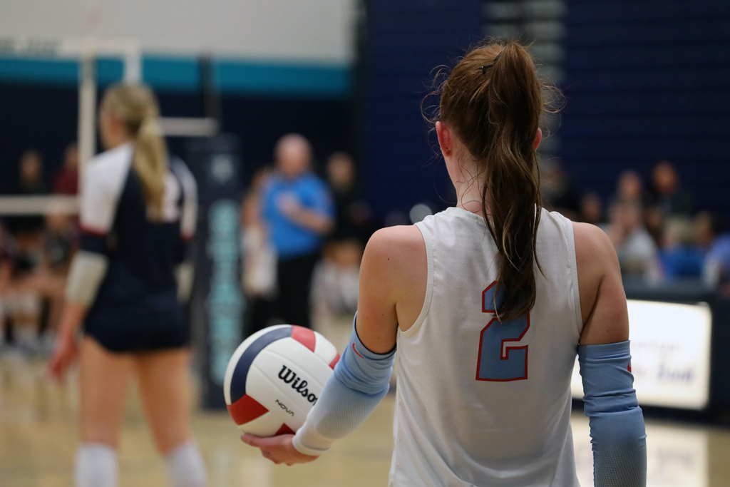Two women wearing sports uniforms stand on a court. One holds a volleyball and faces away.