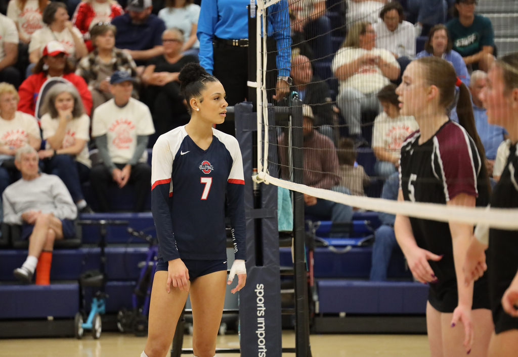 Two volleyball players on court, one watching, others sitting in bleachers, volleyball net between them.