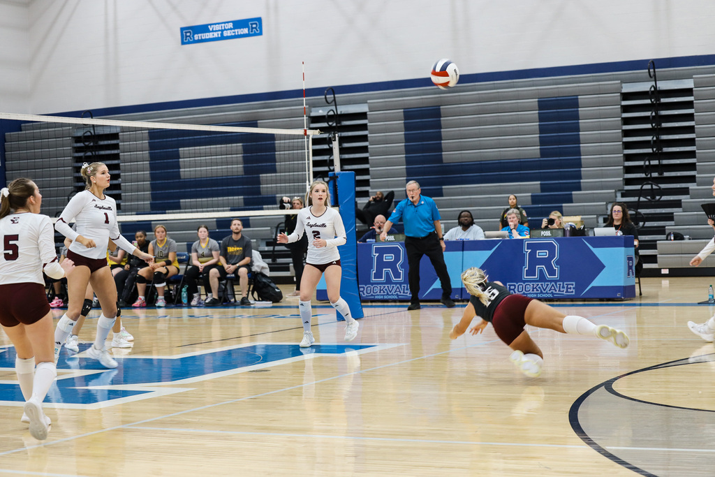 Players in white and maroon uniforms playing volleyball on court; one player diving for ball.